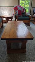 Solid wood farmhouse style coffee table with boarded top and thick wood base shown from a low angle in living room setting with armchair nearby.