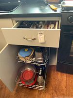 Open drawer showing various kitchen utensils including measuring spoons, spatulas, ice cream scoops, pastry blender, and scissors. Under drawer wire shelving holds storage containers and metal pots.