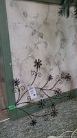 Copper colored metal flower wall hanging leaning against a green and floral wallpapered wall, showing detail of flower shapes and vine patterns.