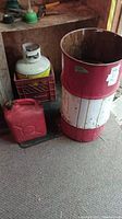 Photo showing the propane tank inside a red milk crate, the metal drum, and the vintage red gas can on carpeted floor.