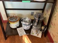 Photo showing the lot of three items on a dark shelf: a Crock-Pot slow cooker, Betty Crocker rice cooker and food steamer, and Cuisinart food processor with manuals and original box for food processor.