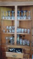 View of three wooden shelves in a cabinet filled with various types of glassware including blue stem wine glasses, clear wine glasses, shooter glasses, martini glasses and Kentucky Derby themed glasses. Drawer below the shelves is open showing assorted flatware.