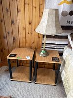Two small end tables with light wood tops and black legs; one table top worn. Brass swing arm table lamp with beige fabric lampshade placed on right table.