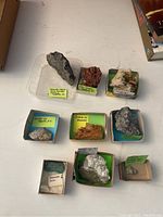Top view of nine mineral specimens in small boxes with handwritten labels showing their names and origins.