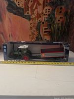 Front view of boxed Bruder Fendt 209 S tractor with trailer showing entire set horizontally with background of a painted wall and ruler for length measurement.