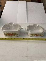Two Corning Ware cooking dishes of different sizes with clear glass lids and Spice of Life vegetable pattern on sides.