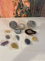 Two rough geode halves, one polished geode slab, and seven small cut stones in plastic bags arranged on a white surface with a colorful painting backdrop.
