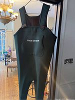 Front view of green Field & Stream chest-high fishing waders with logo on chest pocket hung on a hanger indoors.