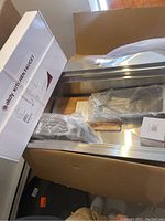 Opened box showing stainless steel kitchen sink with some protective packaging and a dented spot visible, also showing contained items wrapped in plastic.