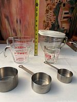 Photo showing two glass Pyrex measuring cups, one plastic Pampered Chef measuring cup with lid, and three stainless steel measuring cups of different sizes
