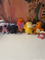 Five plush dolls lined up on a surface: three LOL Surprise dolls on left, two Pokémon plush dolls on right (Psyduck and Charmander).