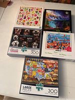Five completed boxed jigsaw puzzles displayed on table: vegetables, landscape, dogs, Paris, USA map.