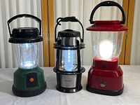 Photo showing all three battery powered camping lanterns powered on and emitting white LED light: green Ozark Trail, black and silver Eddie Bauer, red GE lantern