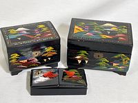 Three black lacquer boxes of varying size placed on a white background, showing colorful hand painted landscapes with trees and mountains.