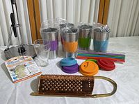 Five stainless steel cups with different colored silicone sleeves, matching lids, colorful straws, Coca-Cola glass, copper grater, SinkShroom packaging visible on white tablecloth.