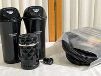 Two black automobile dust bins with push lid and labeled 'Automobile dustbin', black cup holder ashtray with geometric pattern and silver trim, small black 2-port USB car charger outlet, and coiled black desk cable organizer in plastic wrap on white cloth background.