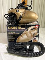 Gold Turbo Tiger handheld vacuum with power cord coiled around it, resting on original branded box, with black hose and attachments displayed in front.
