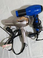 Two hair dryers together, one blue Revlon and one metallic rose gold Conair, cords coiled.