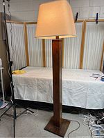 Full view of the tall wood floor lamp showing entire post, base, and shade, in indoor setting against a backdrop with a covered table and divider screens.