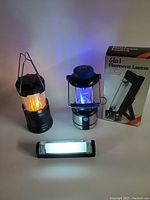 Three battery operated flashlights and lanterns illuminated with colored light effects and white light. One lantern displays orange light, another displays blue light, and the flashlight shows fluorescent white light. Original box for 6in1 fluorescent lantern visible.