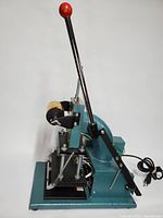 Side front view of the teal cast iron base hot foil stamping machine with chrome lever and red knob, showing adjustable stamping bed and gold foil roller.