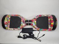 Full view of Hoverheart hoverboard with colorful candy and cartoon pattern, black foot pads, and included charger with power cord layout
