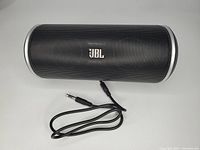 Top view showing black cylindrical JBL Flip speaker with mesh grill and attached audio cable with 3.5mm jack.