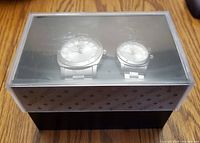 Pair of men's and women's Roots watches inside a black display case with transparent lid. Lid has visible scratches and scuffs.