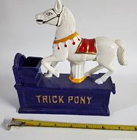 Side view of the white carousel horse figure on the purple base with 'TRICK PONY' lettering alongside a measuring tape showing length about 7 inches.