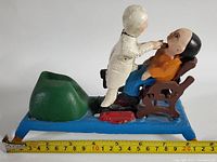Side angle showing dentist pulling tooth from seated patient on blue base with green container, tape measure showing length approx 8.5 inches
