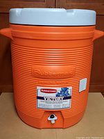 Front view showing Rubbermaid Victory label and spigot on orange cooler with white lid.