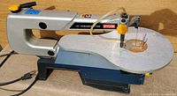 Full view of the Ryobi scroll saw showing silver arm, yellow adjustment knobs, wooden insert in center hole, dust blower tube, and table
