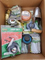 Box containing assorted gardening items including multiple trimmer blades, line spools, and a metal hole digger.