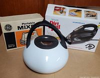Image showing the white whistling kettle with black handle and two boxed appliances behind it (GE mixer and Dirt Devil vacuum).