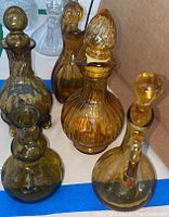 Five amber colored glass oil bottles placed inside a cardboard box with blue tape on the bottom. Bottles vary in shape, size, and design; all have stoppers in place.