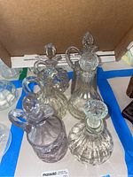 Six assorted clear glass and lead crystal bottles and decanters with stoppers, various styles and patterns, arranged in a box.