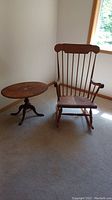View of the oval wooden table with decorative inlay and wooden rocking chair positioned nearby, showing overall condition and style.