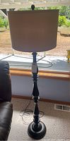 Wooden floor lamp with beige lampshade standing on carpet near window and leather chair