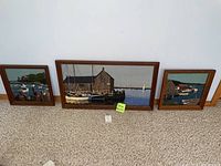 Three framed textile art pieces depicting harbor scenes propped on carpeted floor.