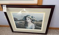 Framed signed art piece showing a young girl with a floral hat, situated in a rural field landscape. Dark wooden frame, cream double mat, glass front.