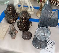 Photo of several ceramic and marble sculptures showing two dark brown busts, an abstract gray marble sculpture, circular marble bowl and a small ceramic oval carving alongside a white relief plaque and larger statues behind.