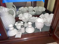 Display of various white milk glass items including hobnail pitcher, salt and pepper shakers, bowls, and footed dishes with ruffled edges in a glass cabinet.
