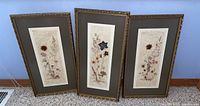 Three framed mixed media floral art pieces leaning against a blue wall on carpet, showing elongated vertical botanical arrangements and decorative frames.