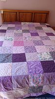 Fully spread purple spring themed quilt on bed with a wooden headboard, patchwork design with various floral and pattern blocks.