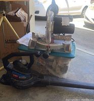 Sears Laser Compound Miter Saw on blue base with angle dial and black motor housing with clear blade guard next to a black Toro Super Blower leaf blower.