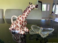 Giraffe ceramic teapot beside clear glass butterfly dish on black surface.