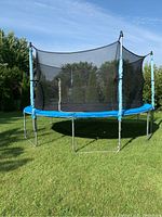 Front-left angle view of the 14ft Jumptek trampoline showing the black mesh enclosure, worn blue pole padding, blue spring padding, and six legs on green grass under sunlight.