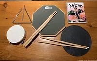 Top view of all items in the lot arranged flat on wood surface, including drum practice pads, drumsticks, triangle, tambourine, and drumming manual.