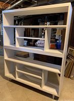 Pair of large white wooden bookcases stacked in a basement or storage area. Shows open shelving arranged in modern geometric compartments. One bookcase has legs and the other stacks on top. Background includes boxes and storage.