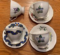 Four fine bone china teacups resting on matching saucers featuring detailed maps and provincial flowers or symbols. Items displayed showing design on cups and saucers.